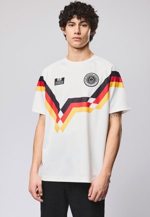 Jonge man met krullend haar, gekleed in een wit sportshirt met rode, zwarte en gele chevronstrepen en logo's op de borst, staand tegen een effen achtergrond.