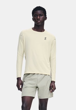 Jeune homme portant une chemise beige clair à manches longues et un short gris clair, debout devant un fond blanc uni.