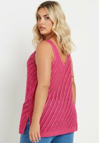 Rosa stickad linne med V-ringning, med ett zickzackmönster, sidoflikar och textilstruktur. Bärs över blå jeans.