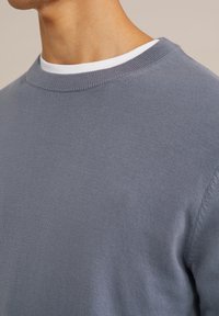 Suéter gris de punto con cuello redondo. Presenta una textura suave y un cuello acanalado. Lleva puesto sobre una camiseta blanca.