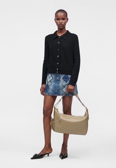 Femme portant un pull noir boutonné et une jupe en jean patchwork, tenant un sac à main beige à courte bandoulière, talons noirs pointus.