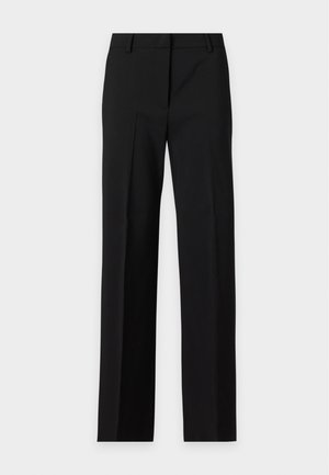 Jeune femme aux cheveux courts et foncés, portant un haut beige sans manches et un pantalon noir à jambes larges, debout devant un fond uni.