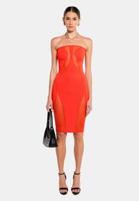 Rotes, trägerloses Kleid mit transparenten Einsätzen, taillierter Silhouette, mittellang und glatter Textur. Kombiniert mit einer schwarzen Handtasche und hohen Schuhen.