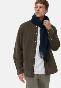 Zielona koszula z długim rękawem i guzikami, wykonana z teksturowanej tkaniny, w połączeniu z granatowym szalikiem z frędzlami na białej koszulce z okrągłym dekoltem oraz jasnoszarymi spodniami.