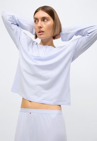 Svetlo modra majica z dolgimi rokavi, okroglim izrezom in zapiranjem na gumbe. Tkanina je gladka in rahlo ohlapna.