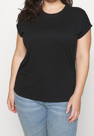 Femme portant un t-shirt noir uni à manches courtes et un jean bleu clair, debout avec sa main droite détendue le long du corps.