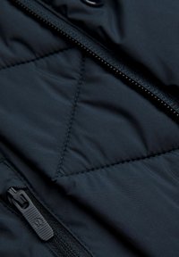 Tissu d'une veste isolée noire avec un motif en losanges, présentant une fermeture éclair latérale, une texture lisse et des coutures piquées.