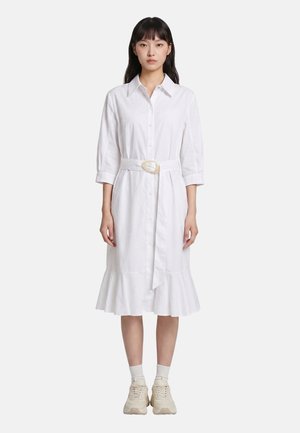 Donna che indossa un vestito bianco con bottoni, cintura, maniche tre quarti e orlo arricciato, abbinato a calzini bianchi e sneakers beige.