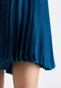 Gonna midi plissettata teal realizzata in tessuto morbido, con pieghe orizzontali e orlo arricciato, che offre una leggera lucentezza e una texture fluida.