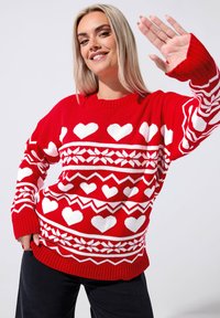 Czerwony sweter z wełny, ozdobiony białymi wzorami w serca, zygzakiem i płatkami śniegu. Ribbowane mankiety i dolna część. Modelka pozuje z uniesioną ręką.