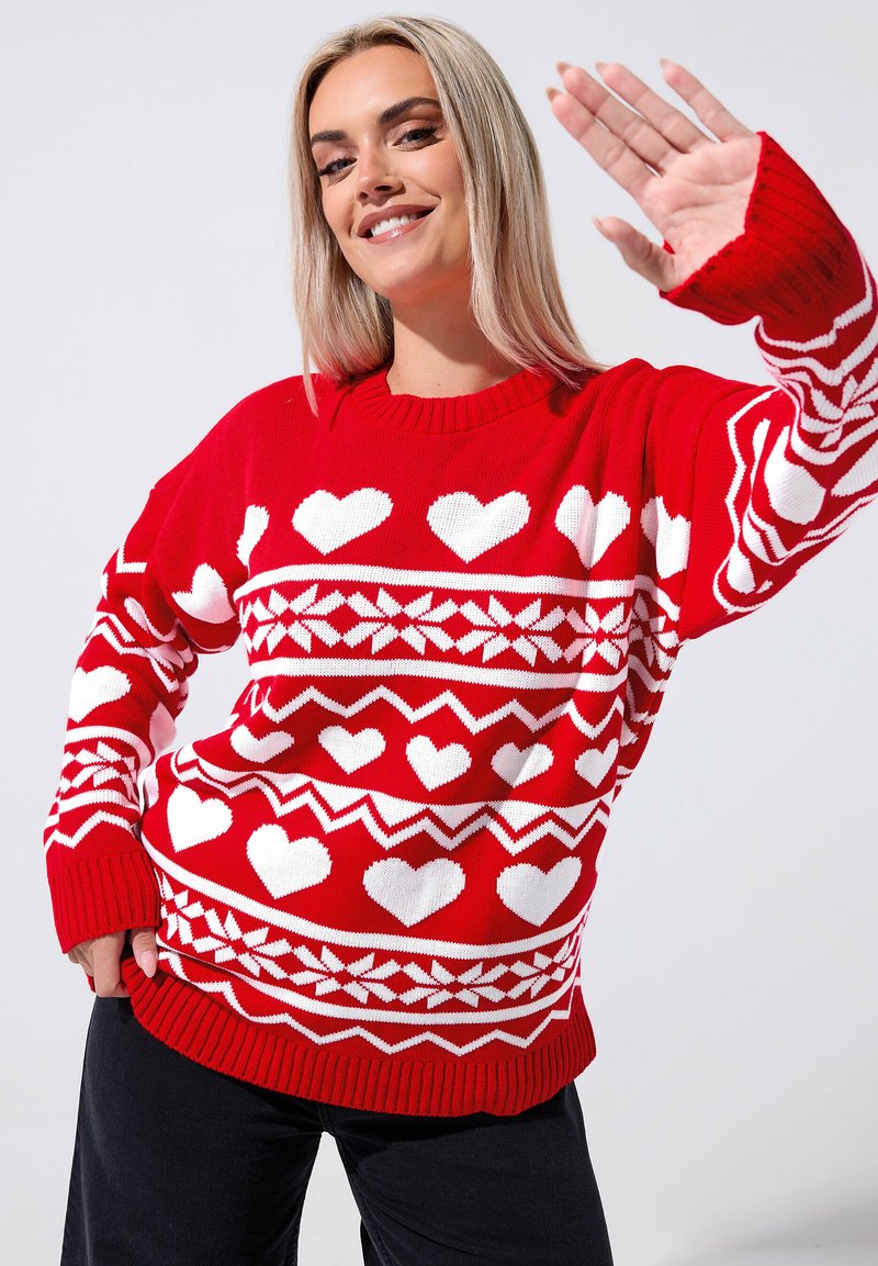 Czerwony sweter z wełny, ozdobiony białymi wzorami w serca, zygzakiem i płatkami śniegu. Ribbowane mankiety i dolna część. Modelka pozuje z uniesioną ręką.