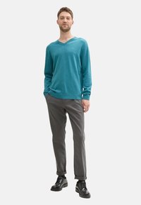 Türkiser V-Ausschnitt-Pullover aus weichem Material, kombiniert mit grauen Hosen. Das Modell trägt schwarze Schuhe mit glattem Lederfinish und Schnürsenkeln.