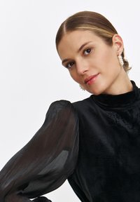 Czarny aksamitny top z przezroczystymi, zwiewnymi rękawami. Posiada wysoki kołnierz i subtelną teksturę. Modelka nosi perłowe kolczyki.