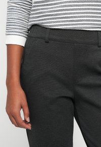 Pessoa vestindo calças cinza escuras e uma camisa de mangas longas listrada em branco e cinza, com a mão esquerda relaxada ao lado.