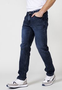 Jeans din denim de culoare albastru închis, cu o croială slim, design cu cinci buzunare și cusături vizibile. Asortați cu pantofi sport albi și gri.