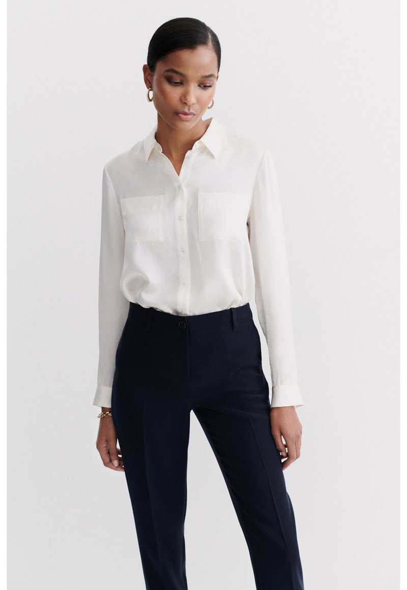 White button-up blouse with long sleeves and two chest pockets, paired with navy tailored trousers. Satin-like fabric with smooth texture.