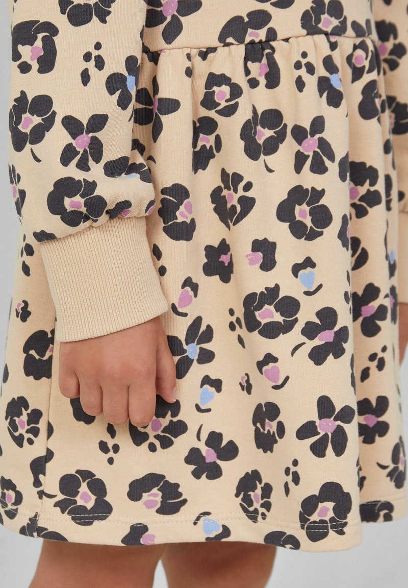 Die Hand eines Kindes ruht an der Seite und trägt ein beiges Kleid mit einem floralem Muster in Schwarz, Lila und Blau sowie einem gerippten Bund an den langen Ärmeln.