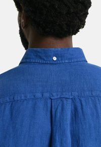 Camisa de lino azul con una costura vertical en la parte posterior, cuello abotonado y un detalle de un solo botón en la parte posterior del cuello.