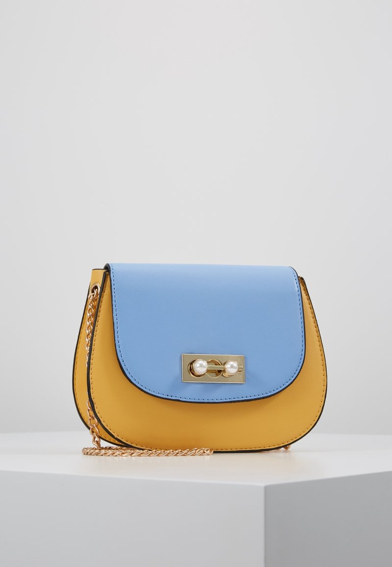 Bolso de mano bicolor con una solapa superior azul y un cuerpo amarillo. Presenta detalles en dorado y un acento de perla en el cierre. Correa de cadena adjunta.