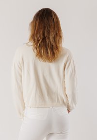 Maglione crema con maniche lunghe, polsini e orlo a coste; indossato con pantaloni bianchi. Morbido al tatto, vestibilità corta, finitura liscia.