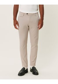 Beżowe, dopasowane spodnie o wąskim kroju, wykonane z gładkiej tkaniny, z przednimi kieszeniami i zapięciem na guzik. Noszone z czarnymi mokasynami i białymi skarpetkami.