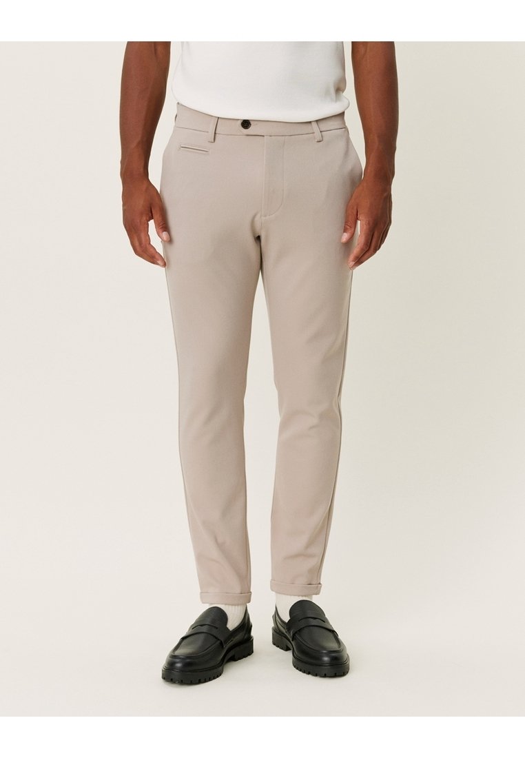 Beżowe, dopasowane spodnie o wąskim kroju, wykonane z gładkiej tkaniny, z przednimi kieszeniami i zapięciem na guzik. Noszone z czarnymi mokasynami i białymi skarpetkami.