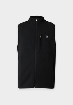 Junger Mann trägt schwarze ärmellose Weste, grauen Pullover mit Bär-Grafik, beige Cargohosen und weiße Sneakers und steht vor weißem Hintergrund.