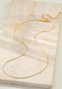 Collier en or avec un design lisse et fin. Comprend un fermoir et une chaîne prolongée pour l'ajustabilité, reposant sur une surface en pierre texturée.