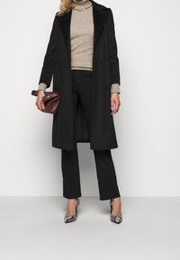 Schwarzer langer Mantel mit Reverskragen, beiger Rollkragenpullover, schwarze Hose und spitz zulaufende High Heels mit Schlangenprint. Hält eine kastanienbraune Clutch.