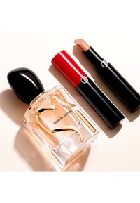 Giorgio Armani parfumflesje met zwarte dop naast rode en beige lippenstiften met zwarte huls op een lichte achtergrond.