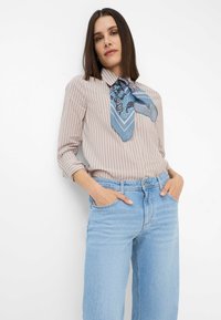 Blusa beige a righe con una sciarpa blu a paisley, abbinata a jeans leggeri azzurri a vita alta. Presenta maniche lunghe e tasche laterali.