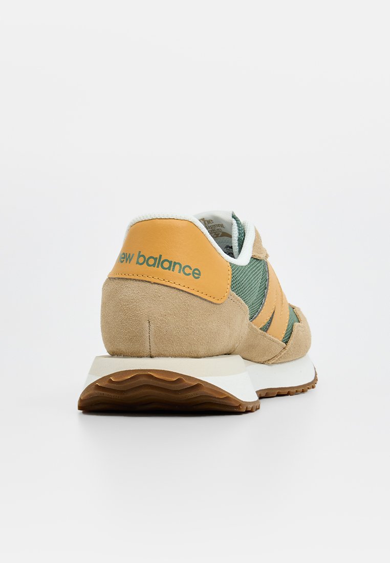 Zapatilla de ante beige y malla verde con acentos naranjas y el logo de "New Balance" en el talón. Suela intermedia blanca y suela de goma.