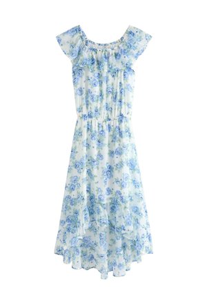 Pige stående med hånden på hoften iført hvid og blå blomsterprintet kjole med rynkede kanter og høj-lav kant samt beige sneakers med hvide ankelsokker.