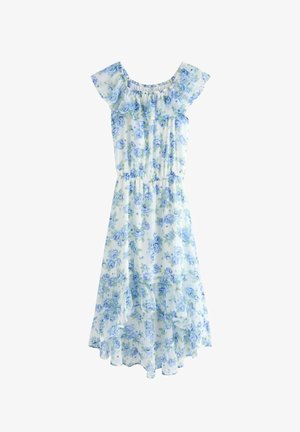 Pige stående med hånden på hoften iført hvid og blå blomsterprintet kjole med rynkede kanter og høj-lav kant samt beige sneakers med hvide ankelsokker.