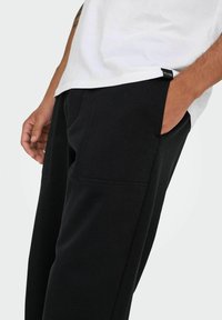 Sorte sweatpants med en glat tekstur, der har to forlommer og kontrasterende syninger. Modellen bærer en hvid T-shirt.