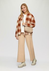 Jongere vrouw in beige wijde broek, crèmekleurige laarzen, witte top, roestkleurige en crèmekleurige geblokte jasje, en beige schoudertas, staand en glimlachend.