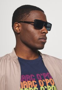 Occhiali da sole rettangolari neri con montatura spessa, indossati con un giubbotto bomber beige chiaro sopra una maglietta blu navy con un design di testo colorato.