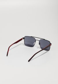 Ochelari de soare cu ramă pătrată, dotate cu lentile gri închise. Podul din metal negru și temple din plastic accentuate cu roșu. Design minimalist.