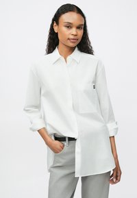 Cămașă albă cu mâneci lungi, cu guler tip button-down, având un buzunar pe piept și un detaliu cu logo. Fabricată dintr-un material ușor, cu o textură netedă.