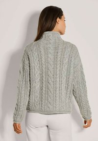 Frau mit langen dunklen Haaren, die einen hellgrauen, strukturierten Pullover und weiße Hosen trägt, steht mit dem Rücken zu einer schlichten, hellen Hintergrund.