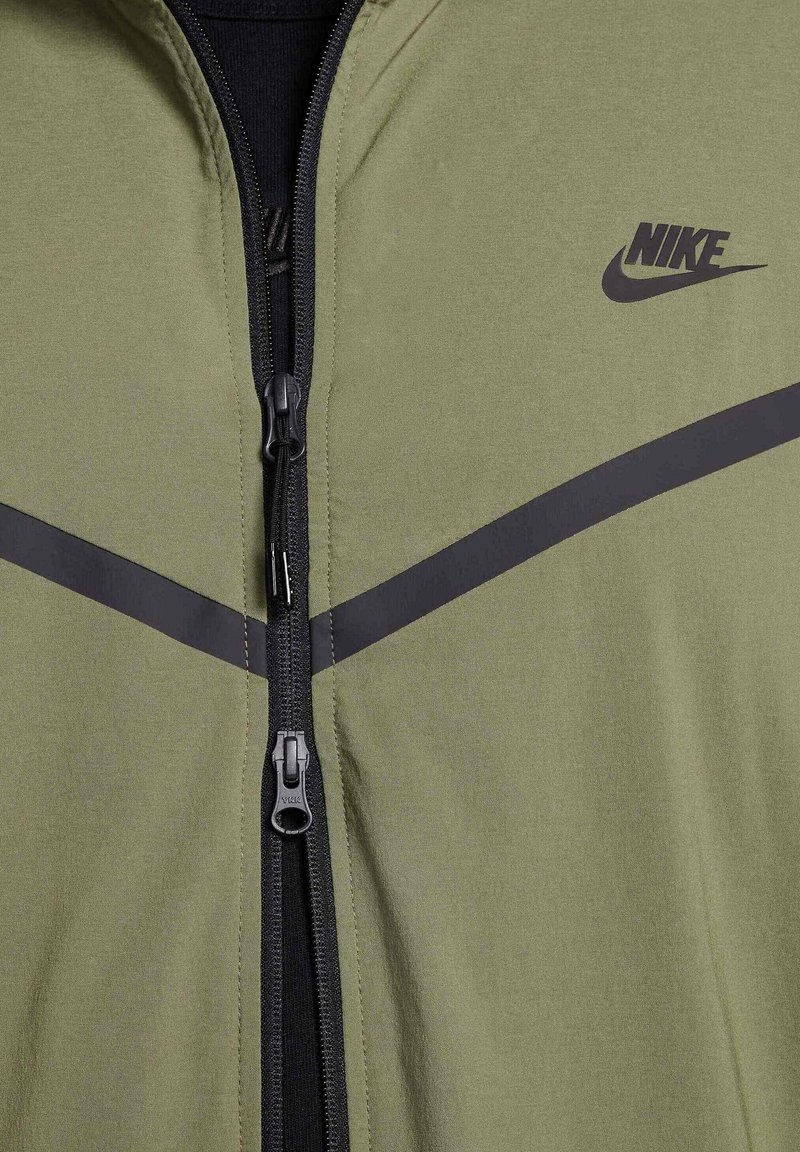 Zielona sportowa kurtka z wysokim kołnierzem, czarnym zamkiem błyskawicznym i poziomą czarną paską na klatce piersiowej; posiada logo Nike na górnej lewej stronie.