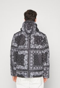 Man in een zwarte hoodie met witte paisley- en bloempatchworkpatronen, die van de camera af kijkt tegen een effen achtergrond.
