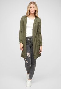 Pitkä oliivinvihreä kardigan, joka on valmistettu pehmeästä kankaasta, avoin etuosa, jossa on kaksi etutaskua, yhdistettynä revittyihin mustiin farkkuihin ja valkoisiin lenkkareihin.