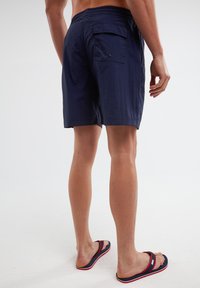 Tamsių jūrų mėlynumo maudymosi šortai su elastinga juosta, užpakaline kišenė ir tekstūruotas audinys, derinami su šlepetėmis jūros mėlynoje, raudonoje ir geltonoje spalvose.