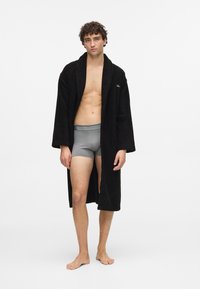 Svart frotté badekåpe med teksturert overflate, åpen front og lange ermer, kombinert med grå tettsittende shorts. Modellen står barbeint mot en hvit bakgrunn.