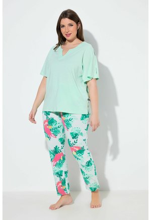 Donna con top a maniche corte verde menta e pantaloni bianchi con stampa di pappagallo rosa e foglie verdi, in piedi a piedi nudi su uno sfondo bianco.