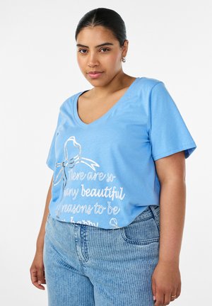 Kobieta ubrana w jasno-niebieski T-shirt z dekoltem w serek z białym napisem "Jest tyle pięknych powodów, by być szczęśliwym" oraz w prążkowane niebieskie jeansy.