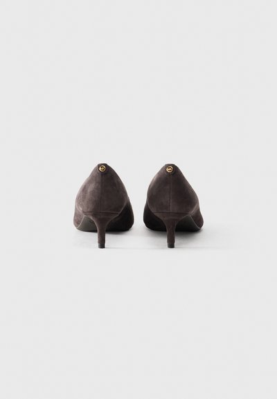 Une paire de chaussures à talons aiguilles en daim marron foncé vue de l'arrière, avec de petits logos circulaires dorés sur la partie supérieure du talon.