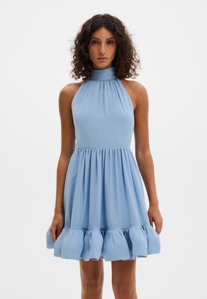 Kvinne med krøllete hår iført ermeløs lyseblå halterkjole med innsvingt midje og volangkant, stående mot hvit bakgrunn.