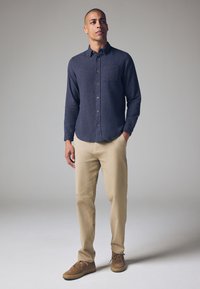 Camisa azul marino de botones con tejido texturizado, bolsillo en el pecho, chinos beiges y zapatos de ante marrón, de pie contra un fondo gris claro.